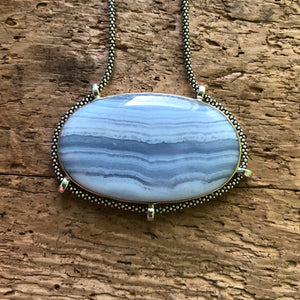 Strikingly good quality horizontally oriented oval Blue Lace Agate Pendant with oxidized sterling silver chain on driftwood background